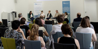 A small lecture hall. Several people are sitting on a stage at the front. Students sit in front of them with their backs to the camera.