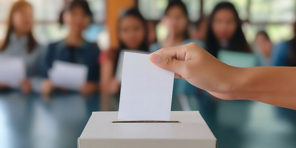 Eine Hand, die einen Wahlzettel in eine Urne wirft. Im Hintergrund sind Kinder zu sehen.